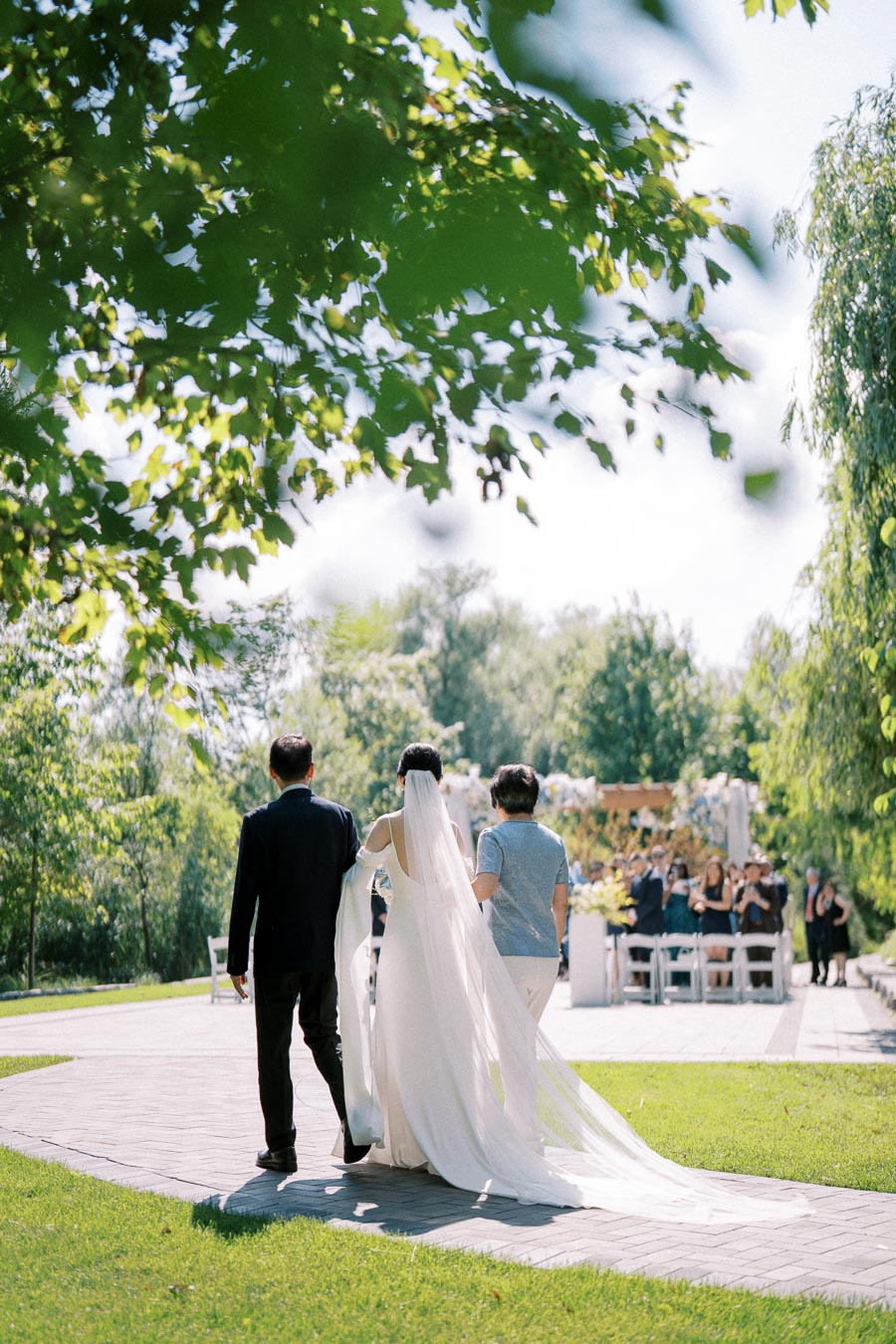 A bride in a flowing white gown walks down an outdoor garden aisle, accompanied by two people, surrounded by lush greenery and sunlight.