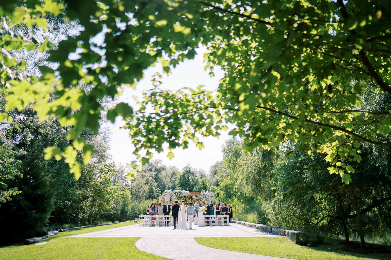 Elegant outdoor wedding ceremony in a lush green garden, featuring the bride and groom standing at an altar surrounded by guests, under a clear blue sky framed by vibrant tree leaves.