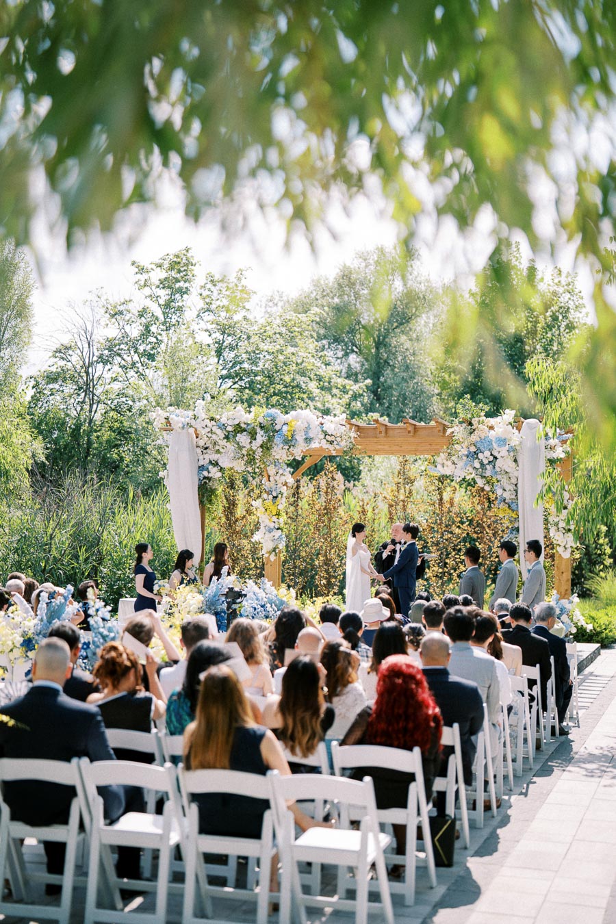 Outdoor wedding ceremony with a couple exchanging vows under a floral arch in a garden setting, surrounded by seated guests.