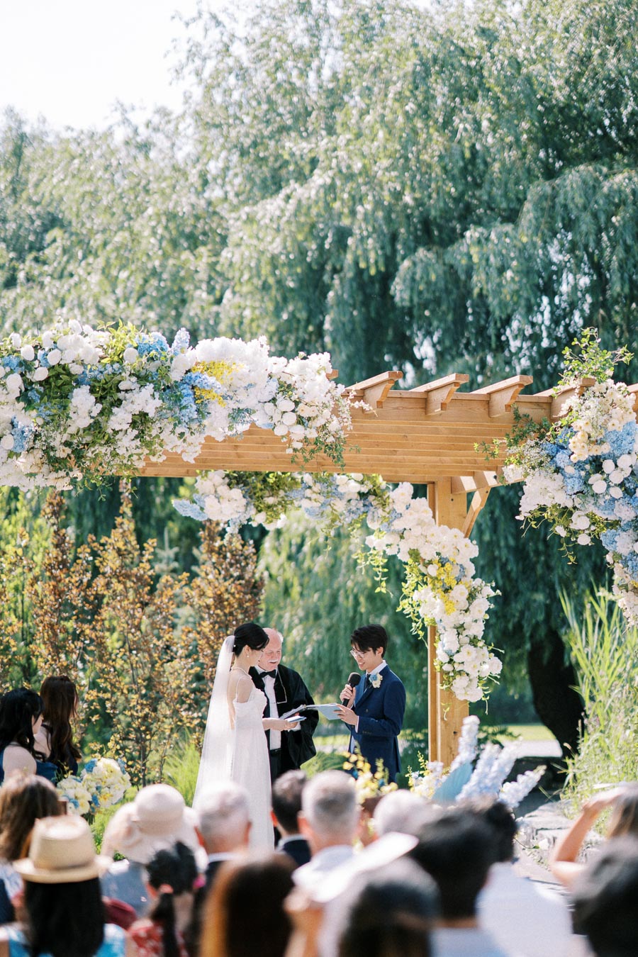 A couple exchanging vows under a wooden pergola adorned with white and blue flowers during an outdoor garden wedding ceremony, surrounded by greenery and guests.