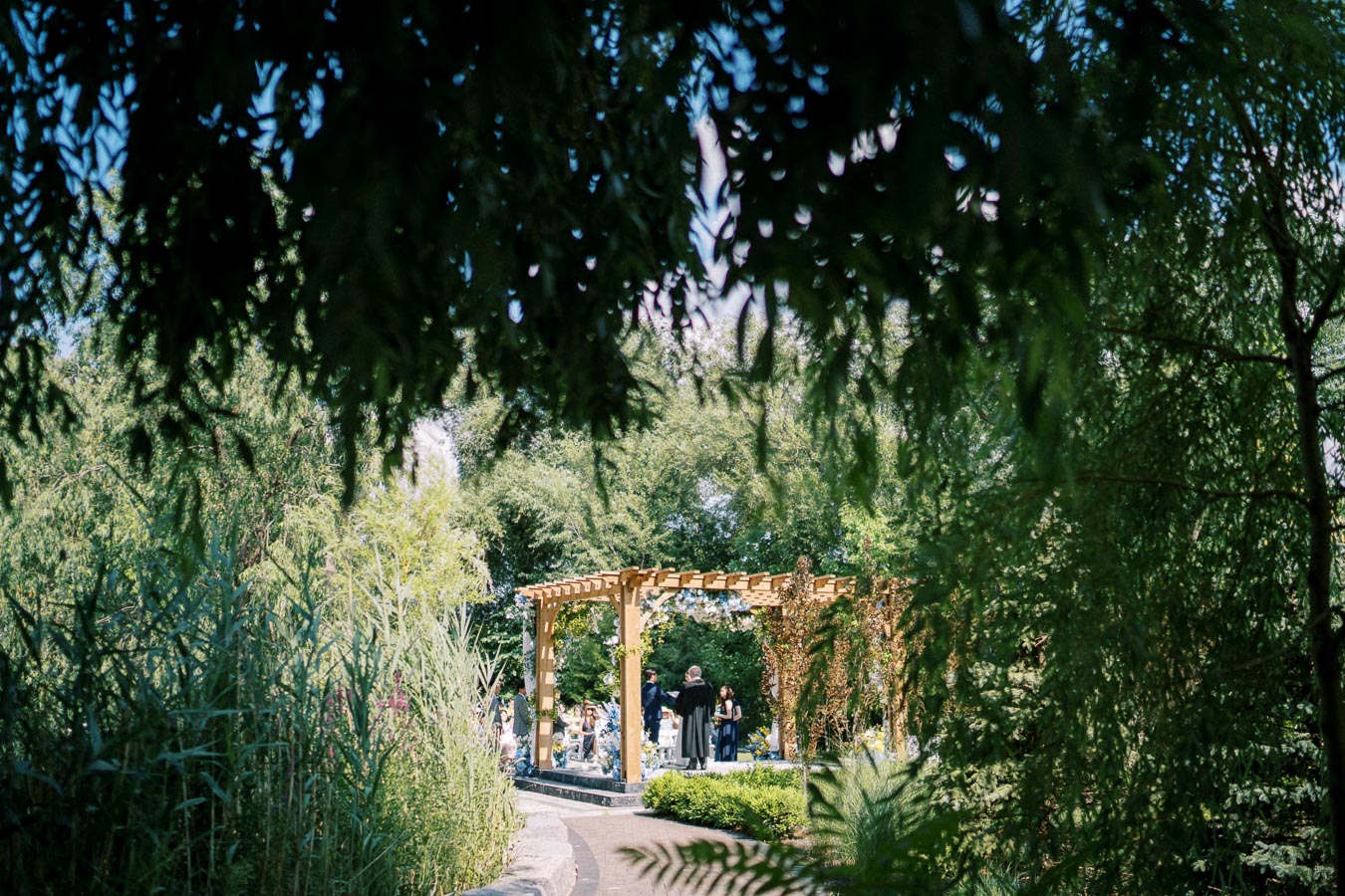 Garden wedding ceremony under wooden pergola, surrounded by lush greenery and trees, with guests seated along paved path.