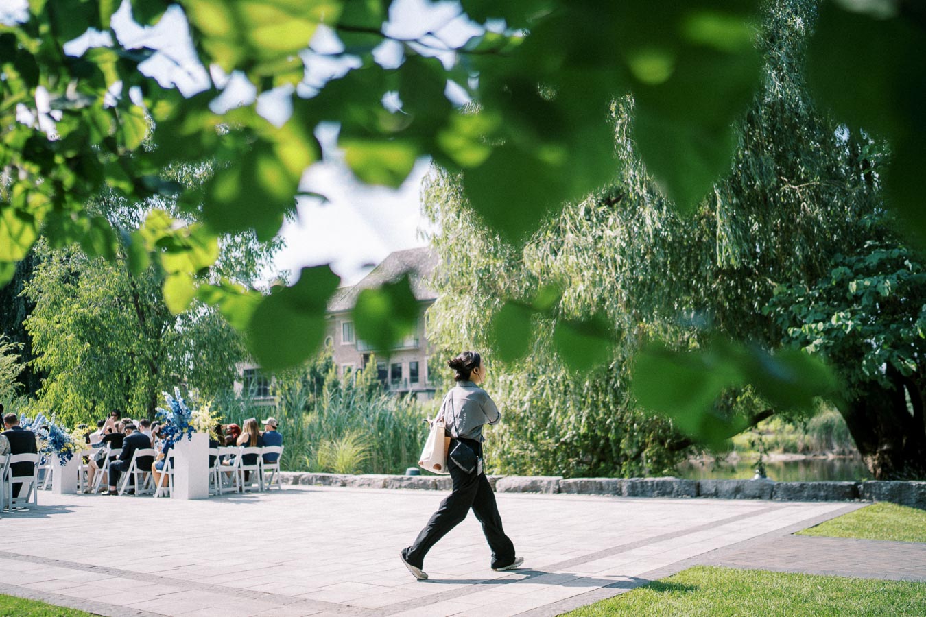 Person walking in a lush garden with a group of people seated at a decorated outdoor event in the background, surrounded by trees and greenery on a sunny day.