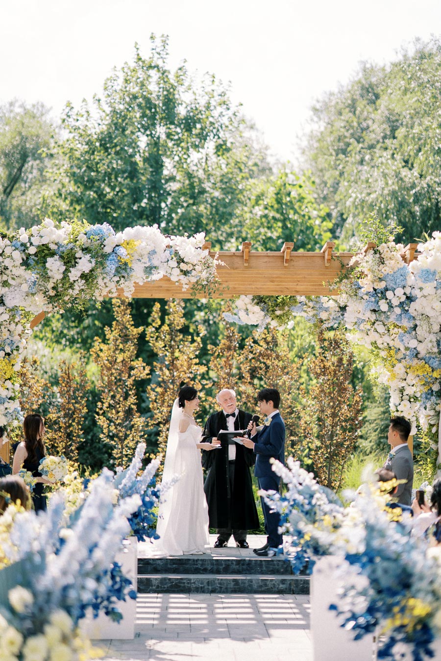 Outdoor wedding ceremony under a wooden arch decorated with white, blue, and yellow flowers, featuring a couple exchanging vows in front of a lush green backdrop.