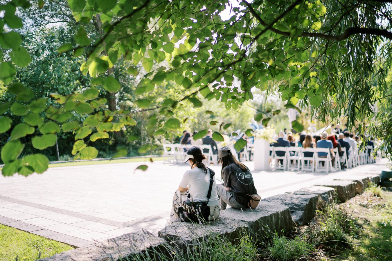 Outdoor wedding ceremony under lush trees, with two guests sitting on a stone ledge in the foreground, and a gathering of seated attendees in the background wearing formal attire.