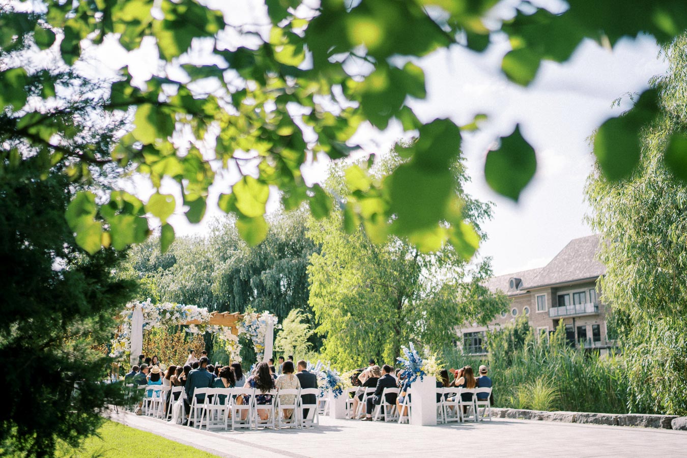 Outdoor wedding ceremony under a floral arbor with guests seated on white chairs, surrounded by lush greenery and trees, under a bright, sunny sky.