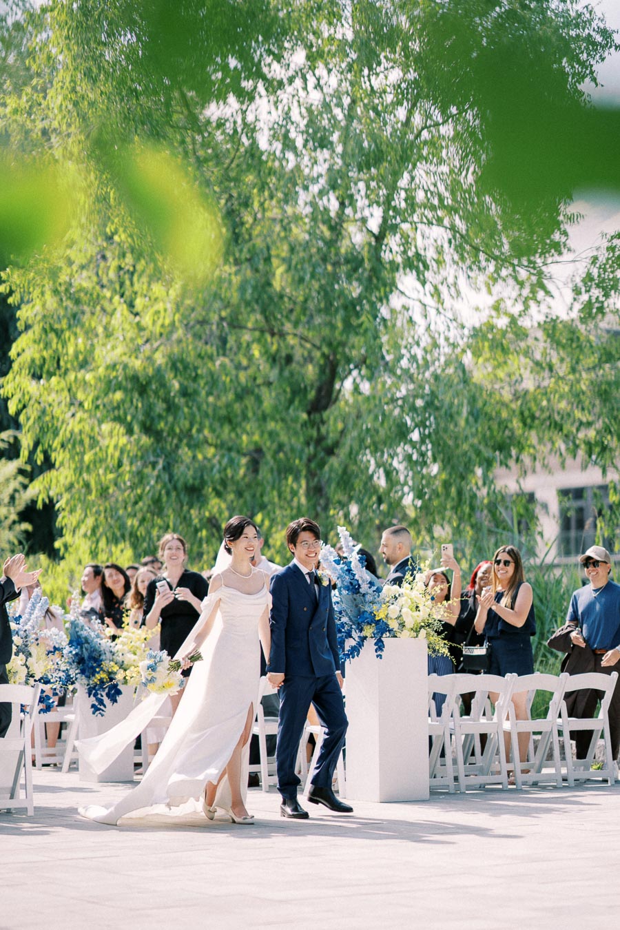 A newlywed couple walks hand in hand down an outdoor aisle adorned with elegant blue and white floral arrangements, surrounded by guests clapping and capturing the moment in a sunlit garden setting.