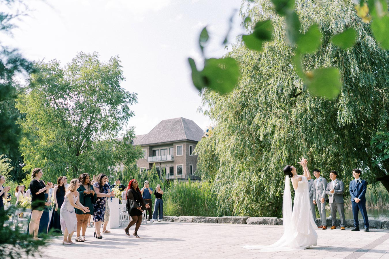 Outdoor wedding scene with a bride in a white dress tossing a bouquet, surrounded by smiling guests eagerly reaching to catch it. Lush greenery and a building are visible in the background, capturing a joyful celebration.