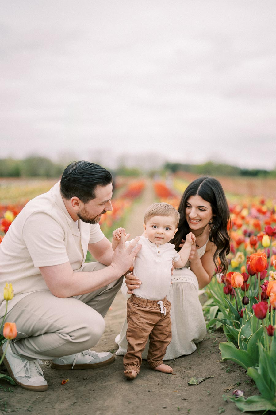 A family enjoying a day in a vibrant tulip field, with the parents supporting their baby as they stand on a dirt path surrounded by colorful flowers.