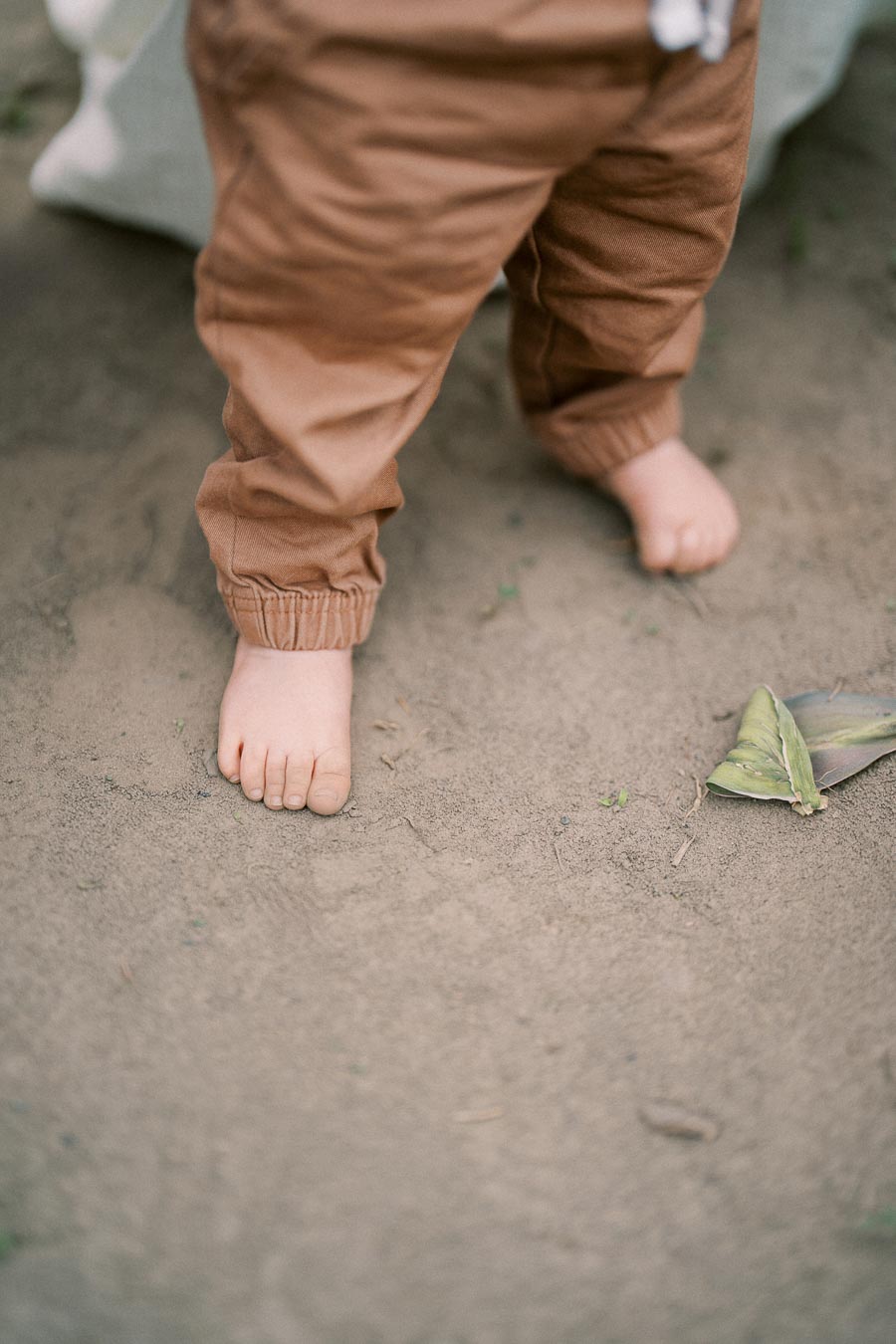 A child stands barefoot on sandy ground, wearing brown pants. A green leaf lies nearby.