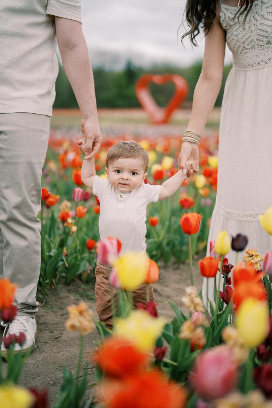 A child stands in a vibrant tulip field, holding hands with two adults, with a heart-shaped sculpture in the background.