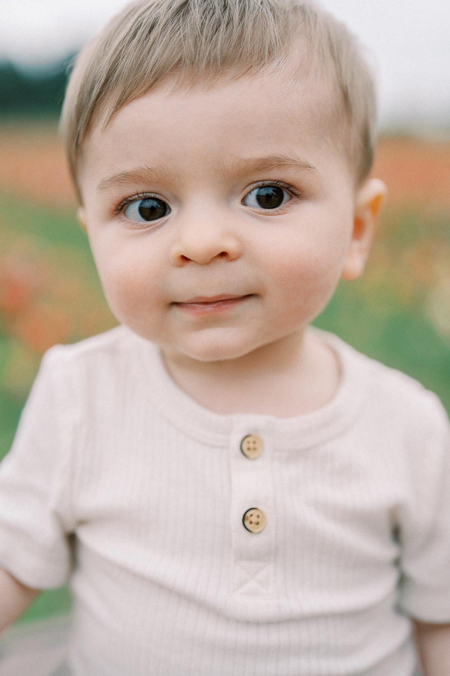 A close-up portrait of a baby with light brown hair and big eyes, wearing a beige shirt with buttons, captured outdoors with a blurred background.