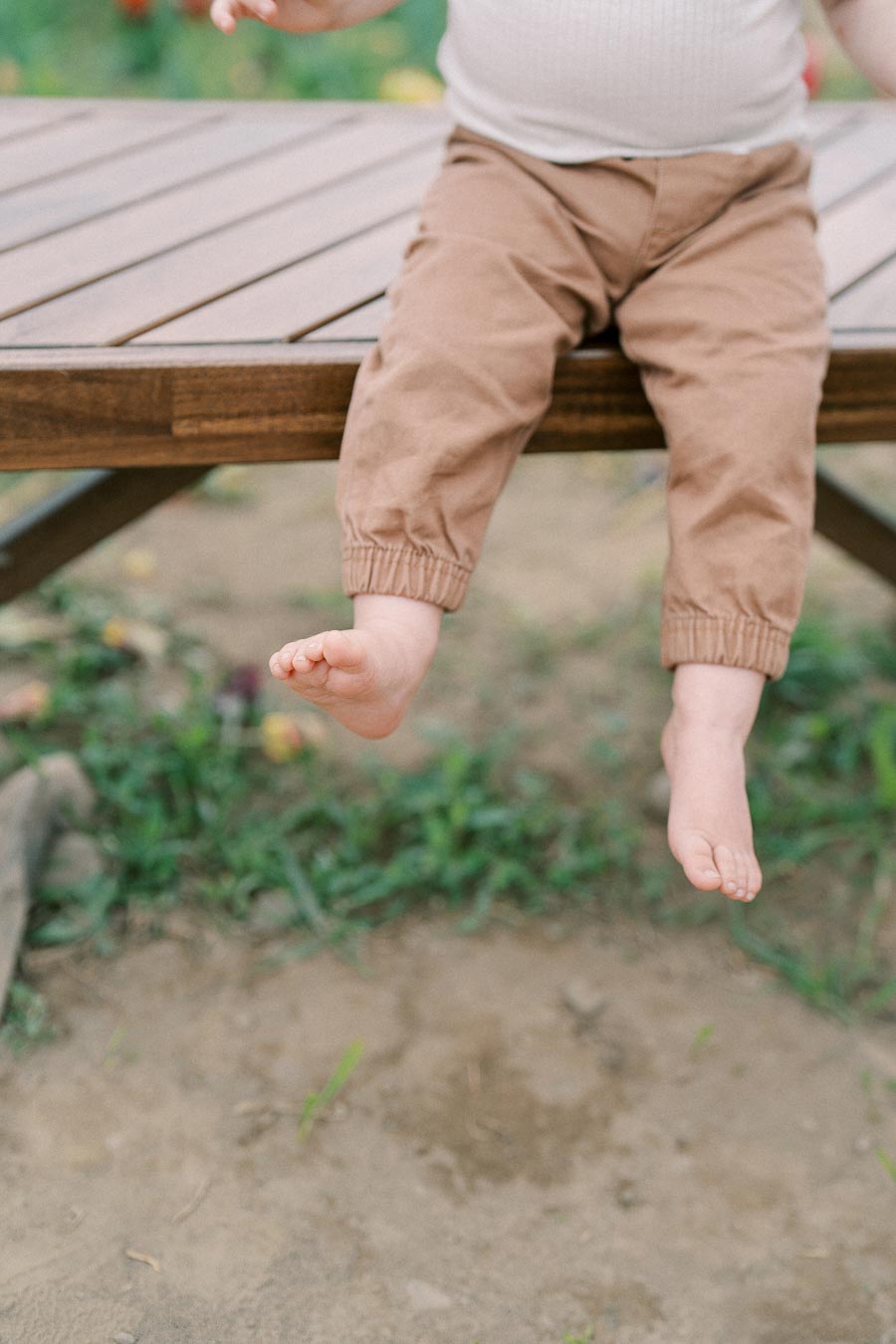 A child sits on a wooden bench with their legs dangling, wearing brown pants and a white shirt.