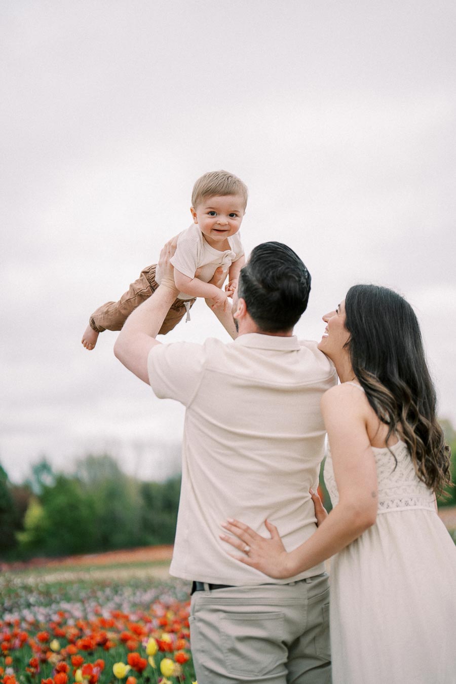 A joyful family moment captured in a vibrant flower field, with a man lifting a baby into the air as a woman smiles beside them.