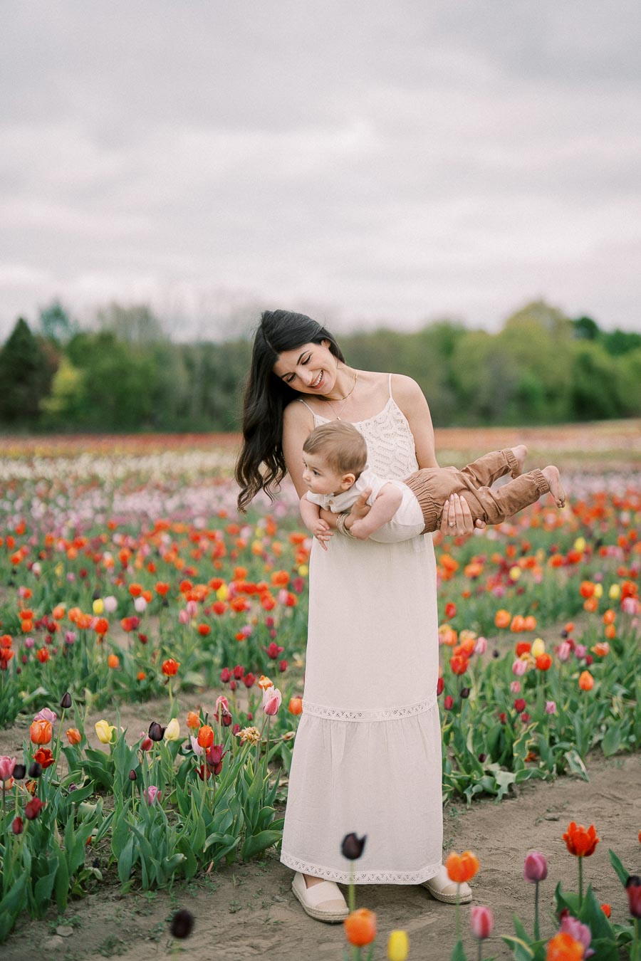 A woman joyfully holds a baby in a vibrant tulip field, surrounded by colorful flowers under a cloudy sky.
