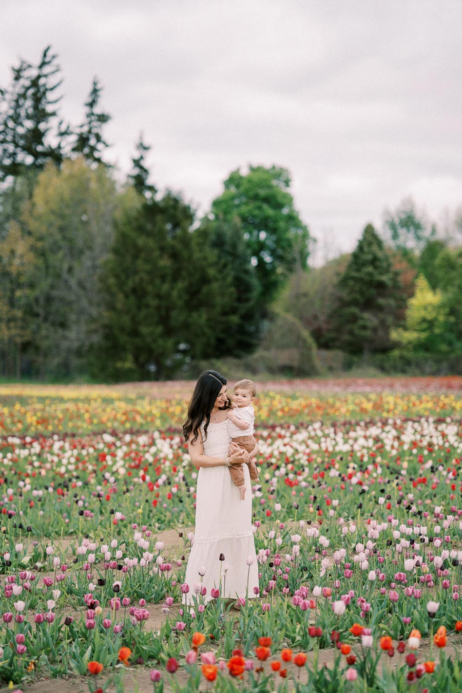 A woman in a white dress holds a baby while standing in a vibrant tulip field, surrounded by a variety of colorful flowers and lush greenery.