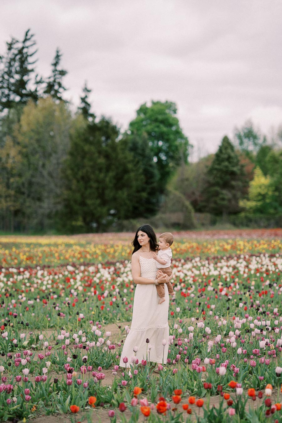 A woman holding a child stands amidst a vibrant field of tulips, surrounded by lush greenery and trees.