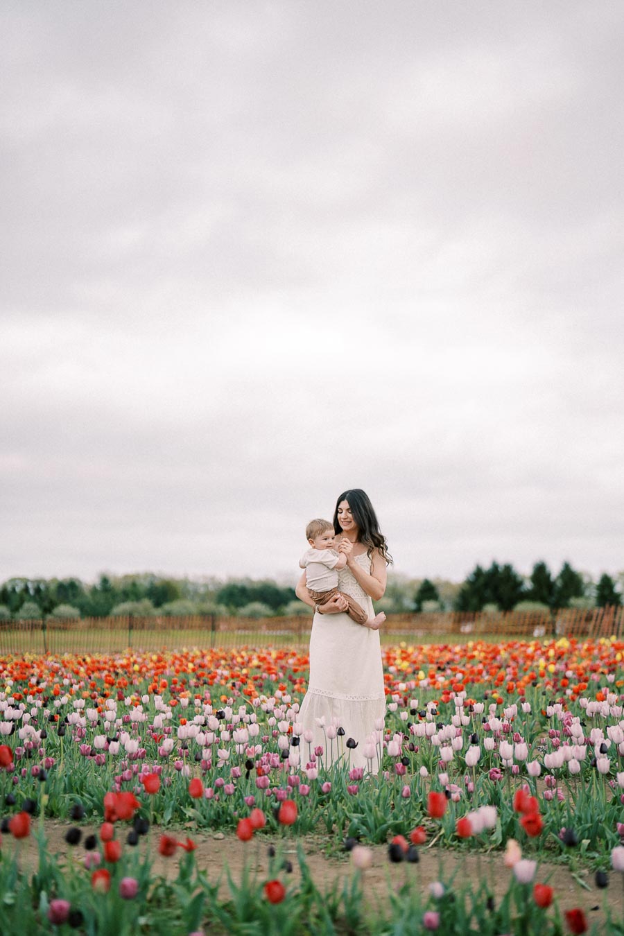 A woman holding a child stands amidst a vibrant field of tulips, showcasing a variety of colors under an overcast sky.
