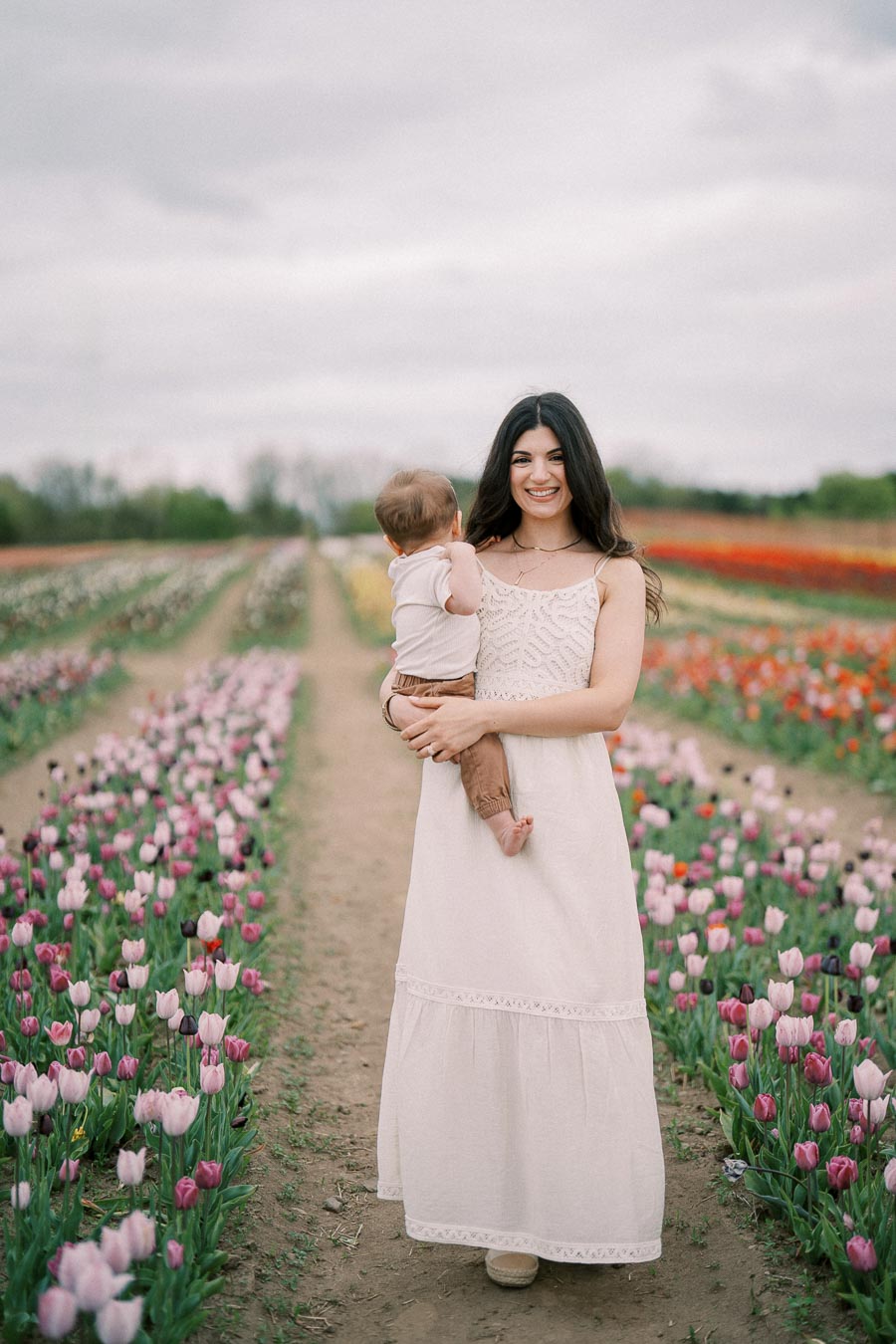 A woman holding a baby stands in a vibrant tulip field, surrounded by rows of colorful flowers.
