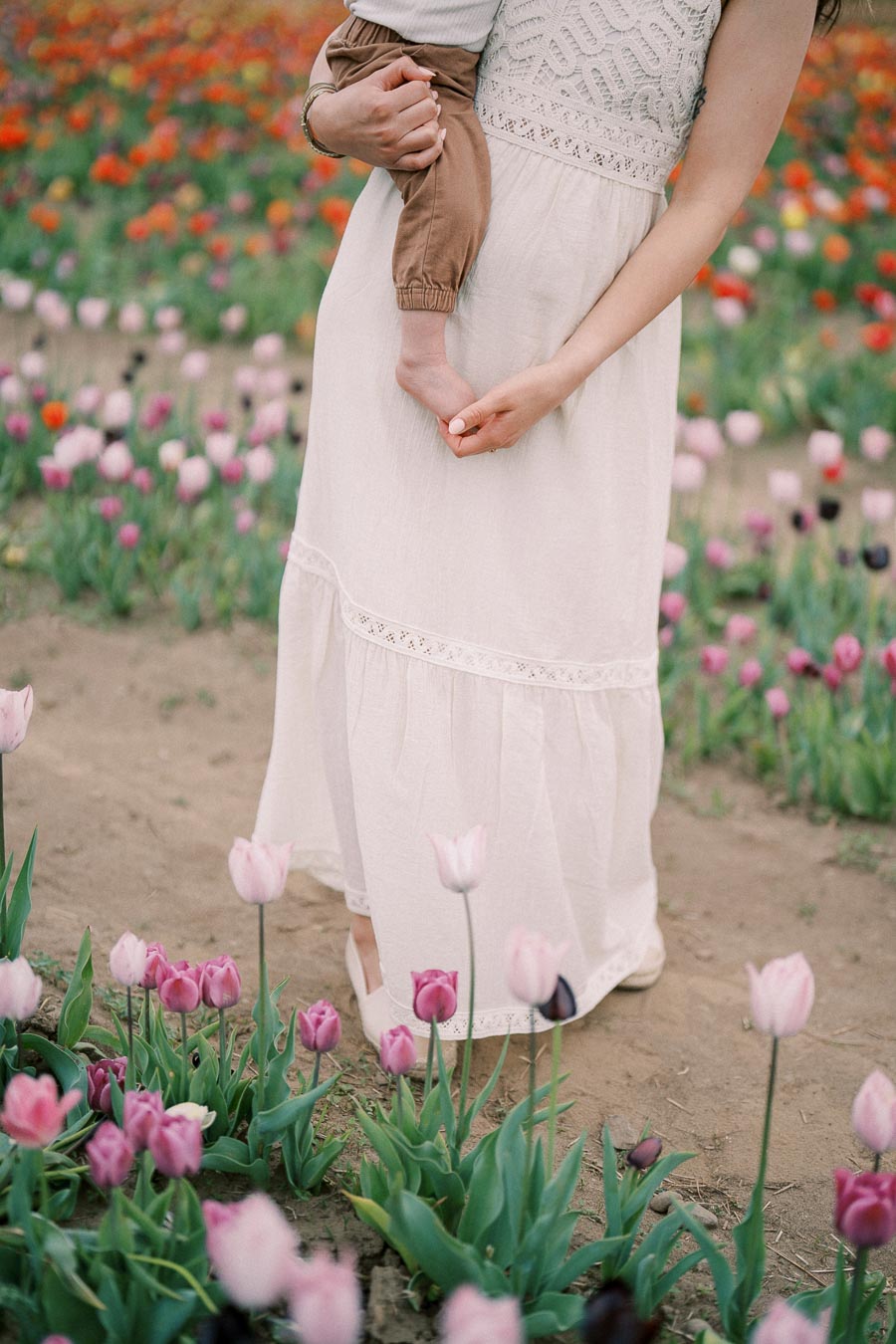 A woman in a white dress holds a child while standing in a vibrant tulip field, showcasing pink and red flowers.