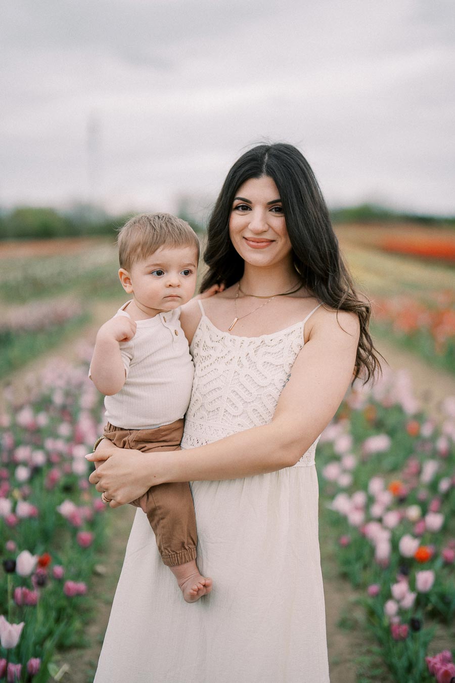 A woman holding a baby stands in a vibrant tulip field, both dressed in light-colored clothing, with rows of colorful flowers in the background.