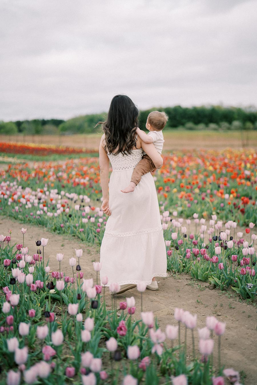 A woman in a white dress carries a baby while walking through a vibrant tulip field, surrounded by pink, purple, and red flowers under a cloudy sky.
