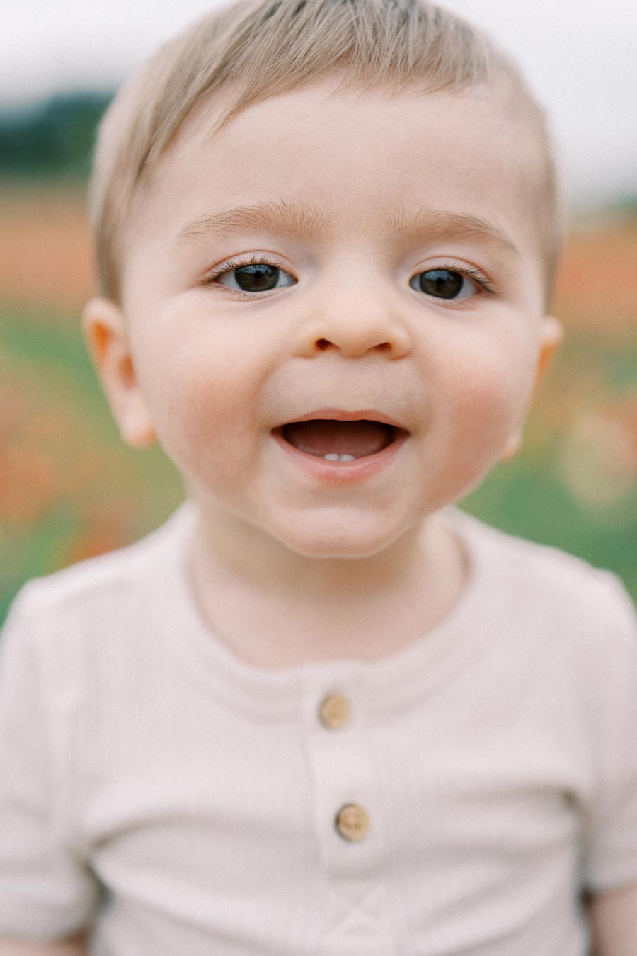 A close-up of a smiling baby wearing a light-colored shirt, captured outdoors with a blurred background.