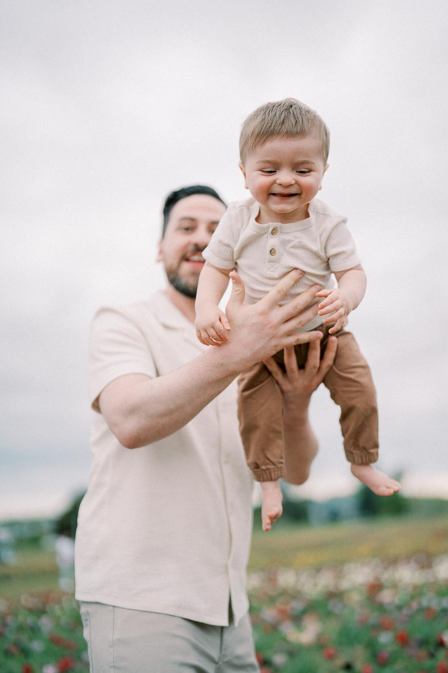 A joyful moment captured as a man lifts a smiling baby into the air in an outdoor setting, surrounded by a field of flowers.