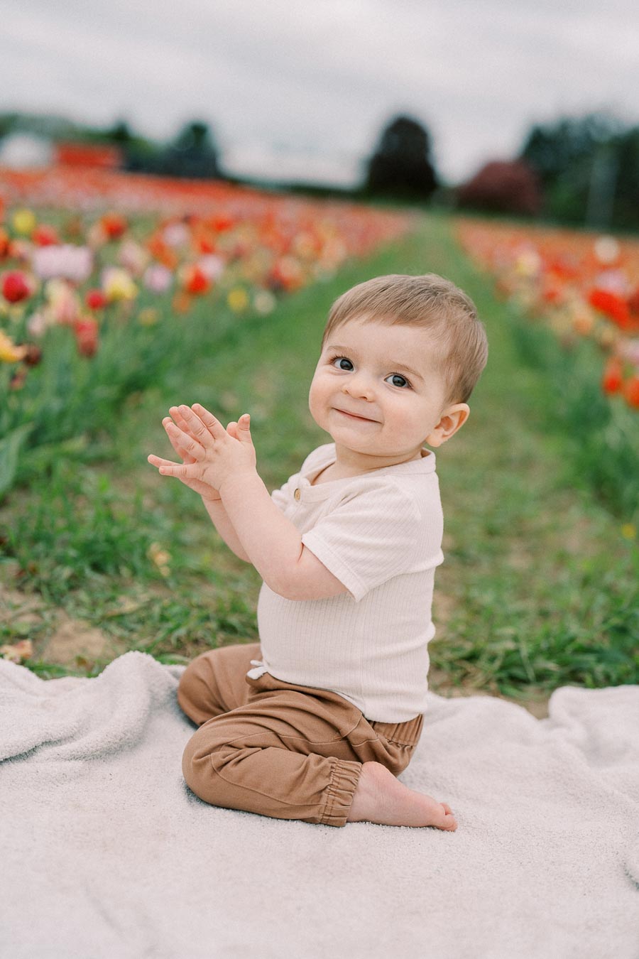 A cheerful baby claps hands while sitting on a blanket in a vibrant tulip field.