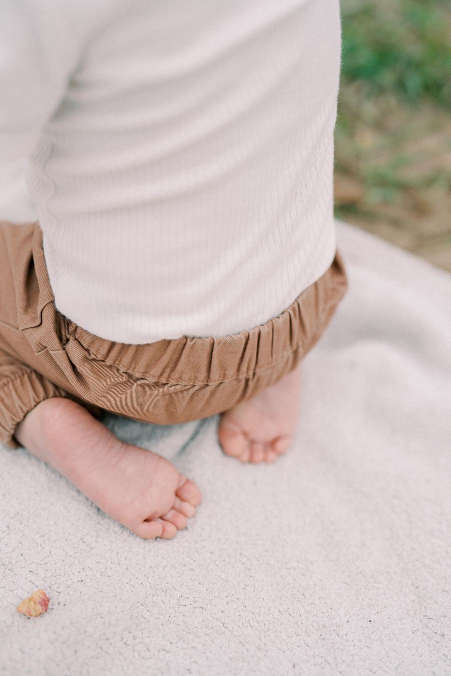 A close-up view of a baby's feet and legs, dressed in brown pants and a white shirt, sitting on a soft blanket outdoors.