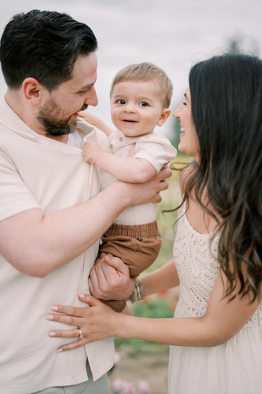 A joyful family moment with a father holding their baby while the mother smiles warmly at them, captured in an outdoor setting.