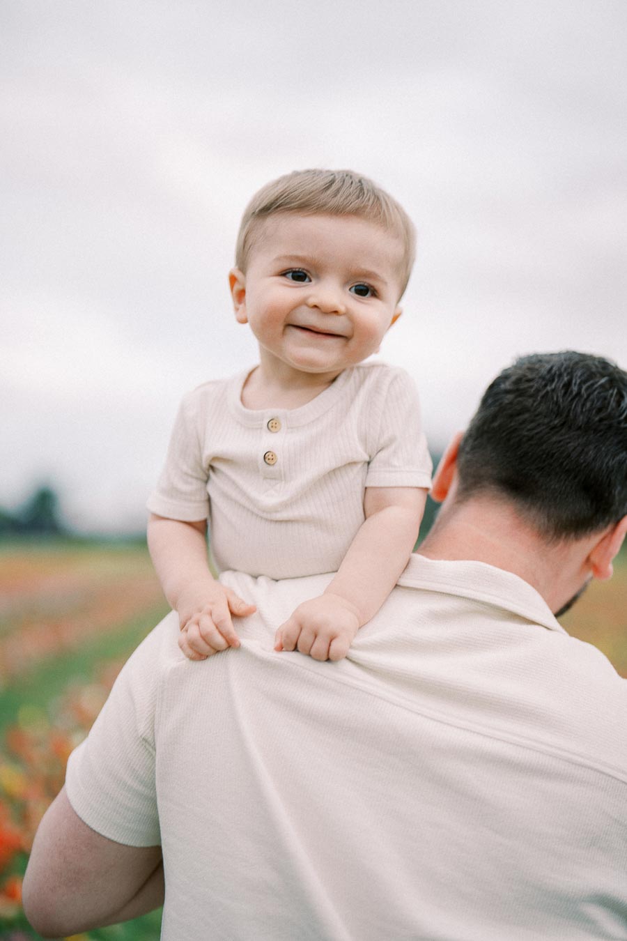 A smiling baby sits on a man's shoulders in a field, both dressed in matching light-colored shirts.