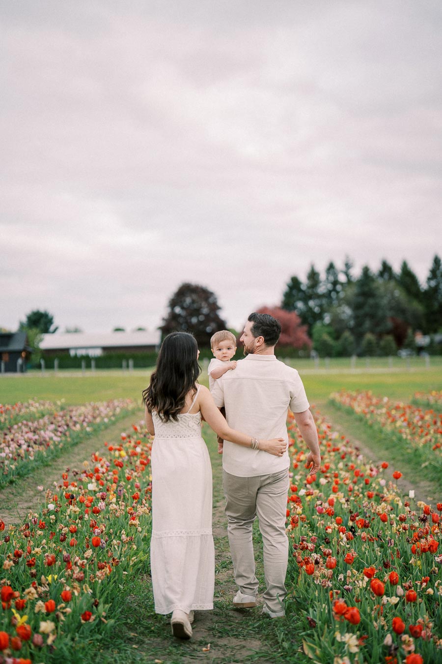 A family strolls through a vibrant tulip field, with the man carrying a child and the woman walking beside him, surrounded by colorful flowers under a cloudy sky.