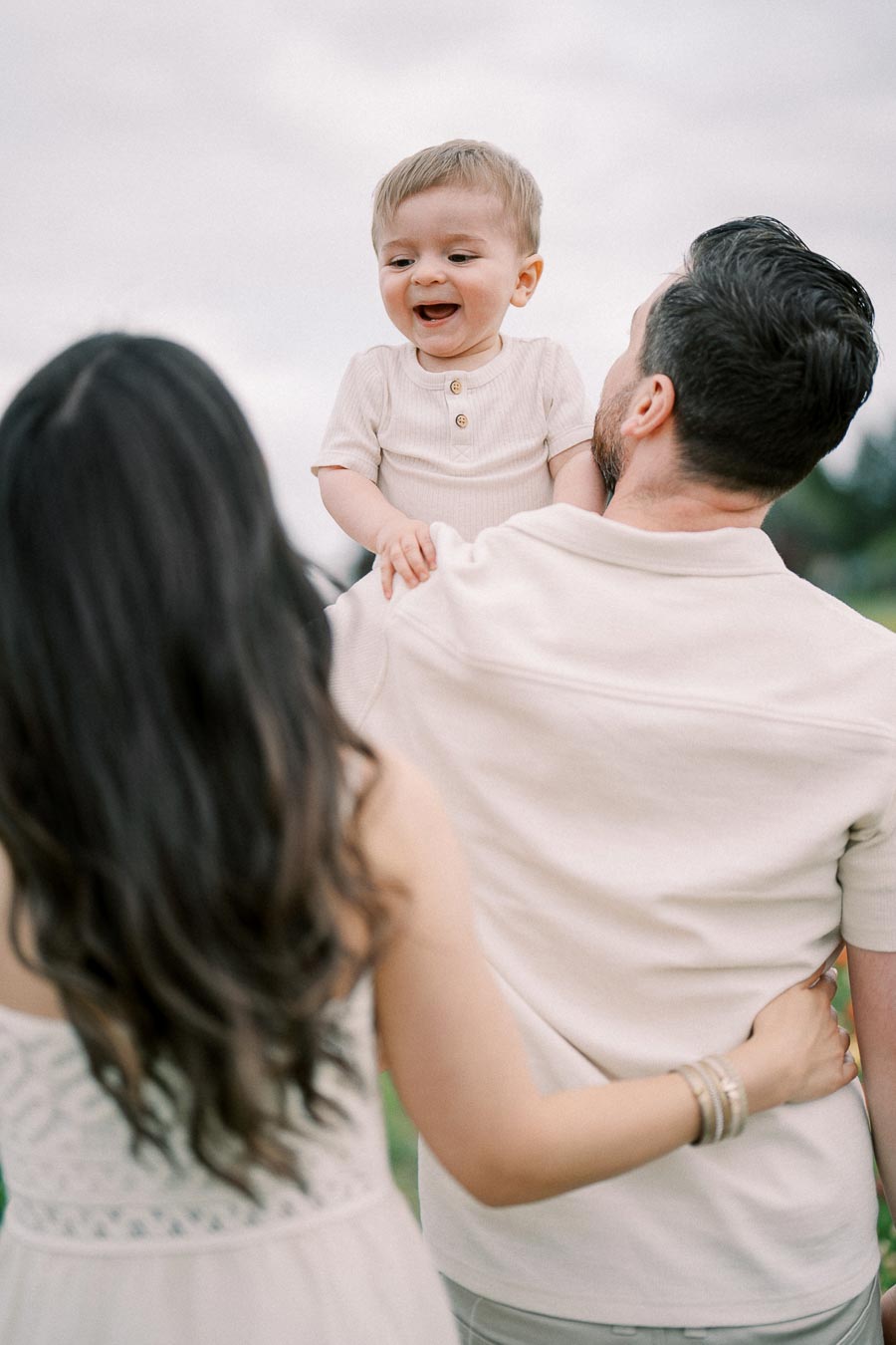 A joyful family moment captured outdoors, featuring a man holding a smiling baby on his shoulder while a woman stands beside them.
