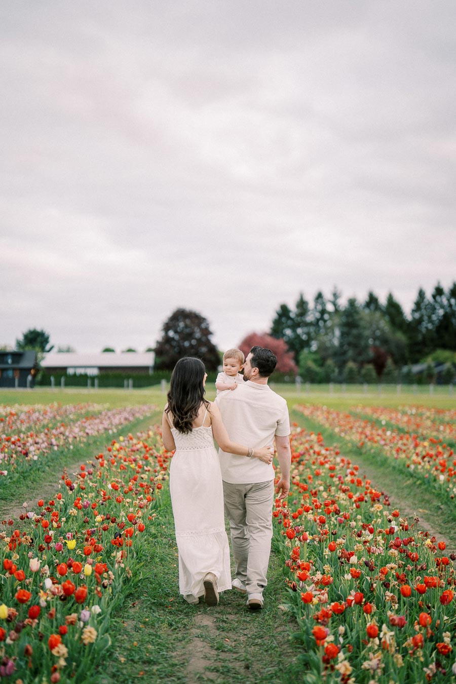 A family strolls through a vibrant tulip field, with the man carrying a child and the woman walking beside him, surrounded by colorful flowers under a cloudy sky.