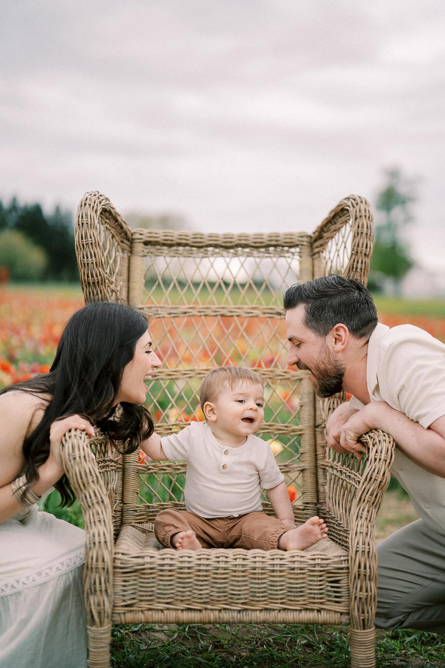 A joyful family moment captured in a field of flowers, with a baby sitting on a wicker chair between two adults.
