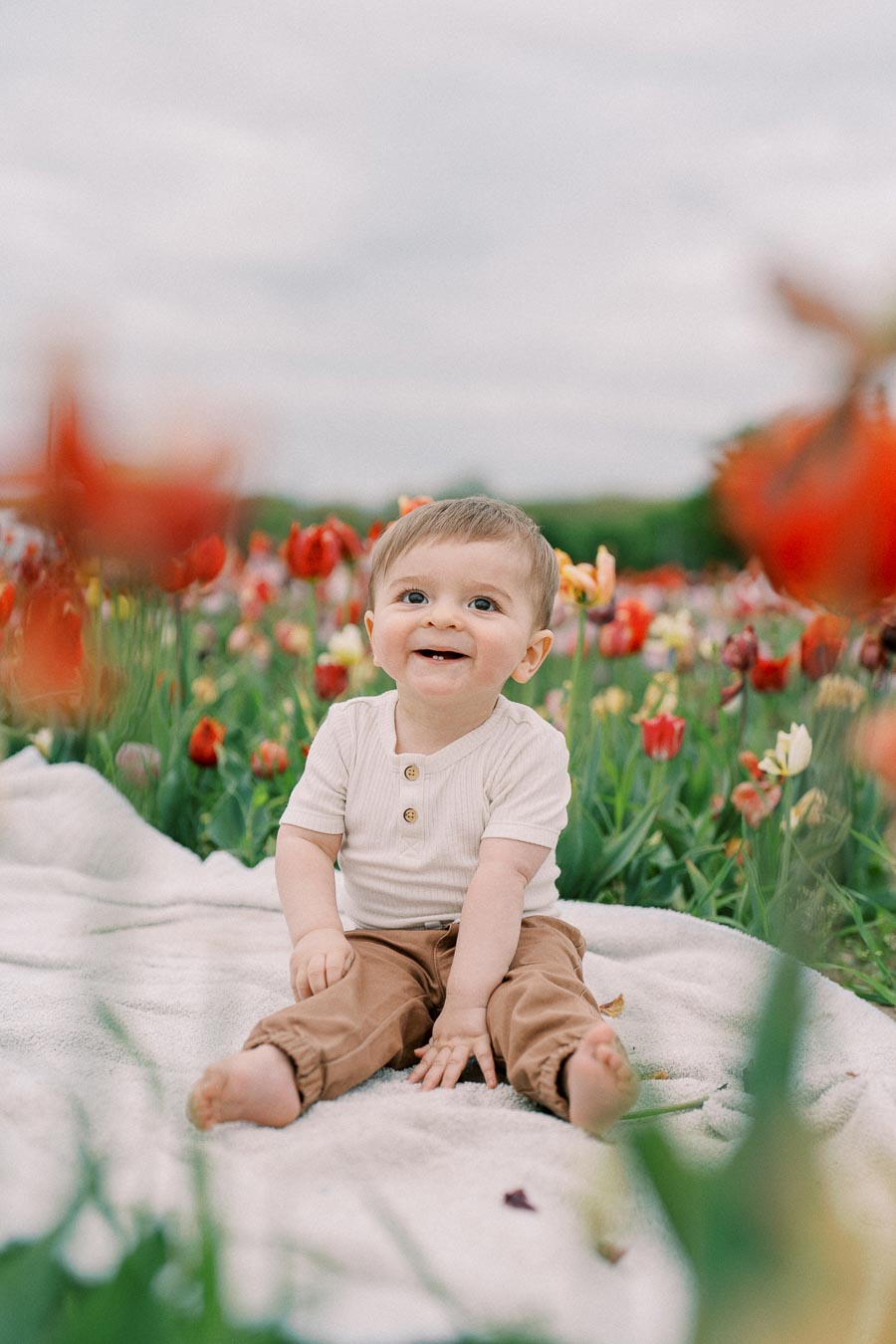 A cheerful baby sits on a blanket amidst a vibrant field of tulips, enjoying a day outdoors.