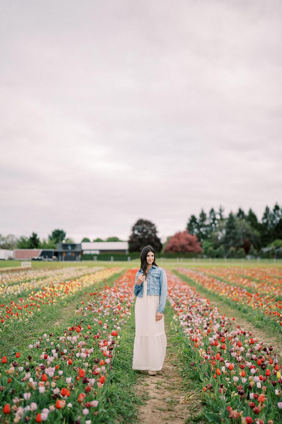 A woman stands in a vibrant tulip field, wearing a white dress and denim jacket, surrounded by rows of colorful flowers under a cloudy sky.