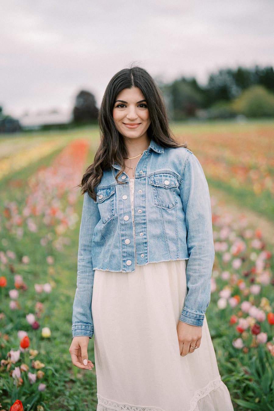 A woman wearing a denim jacket and white dress stands in a vibrant tulip field, smiling at the camera.