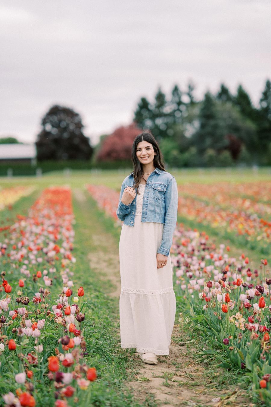 A woman wearing a white dress and denim jacket stands smiling in a colorful tulip field.