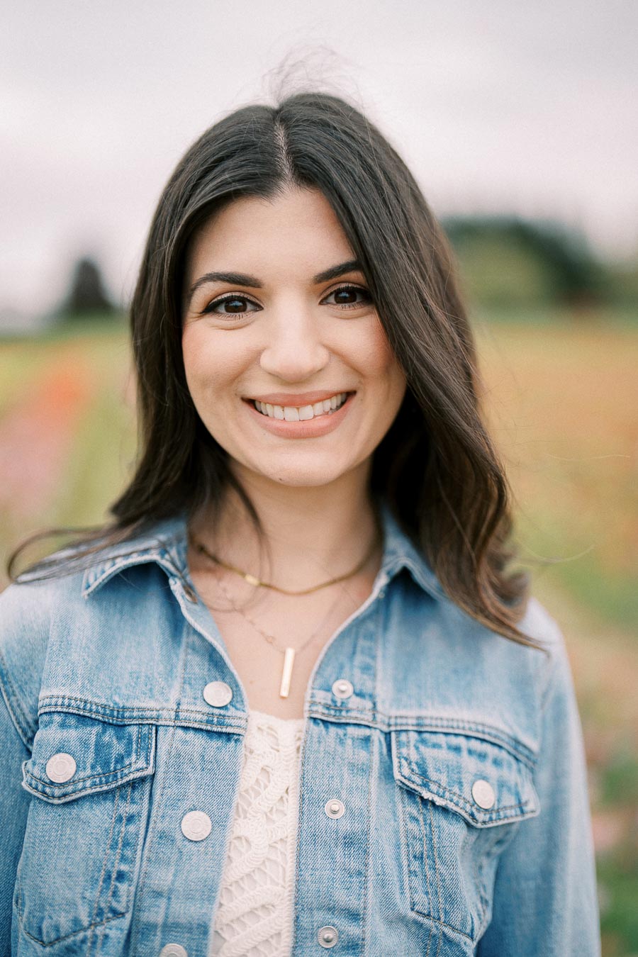 A woman smiling in an outdoor setting, wearing a denim jacket over a white top.