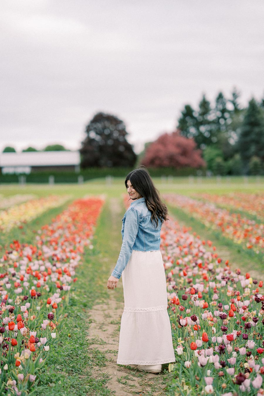 A woman strolls through a vibrant tulip field, wearing a denim jacket and white dress, surrounded by rows of colorful flowers.