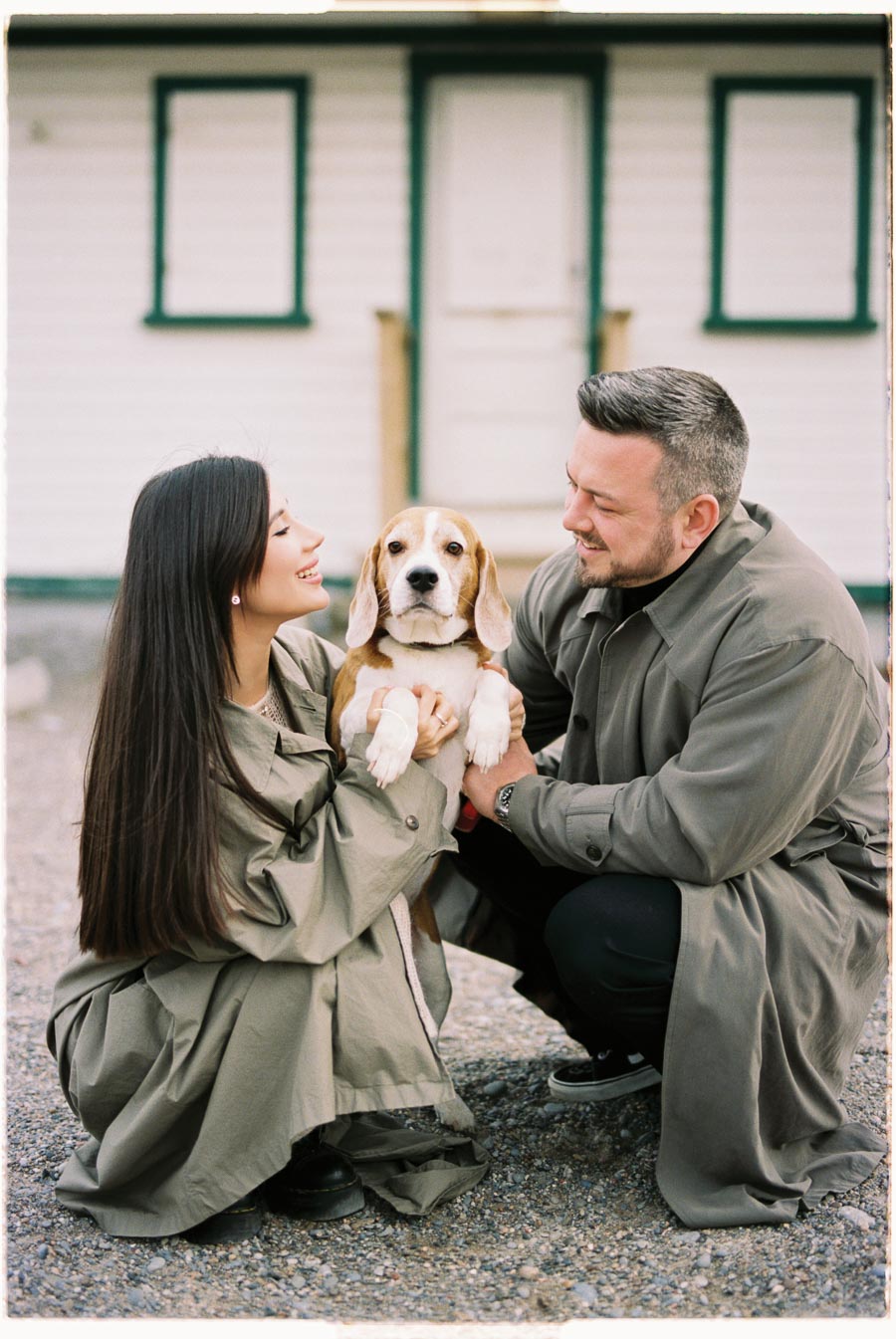 A couple shares a joyful moment with their beagle, dressed in matching trench coats, in front of a quaint building.
