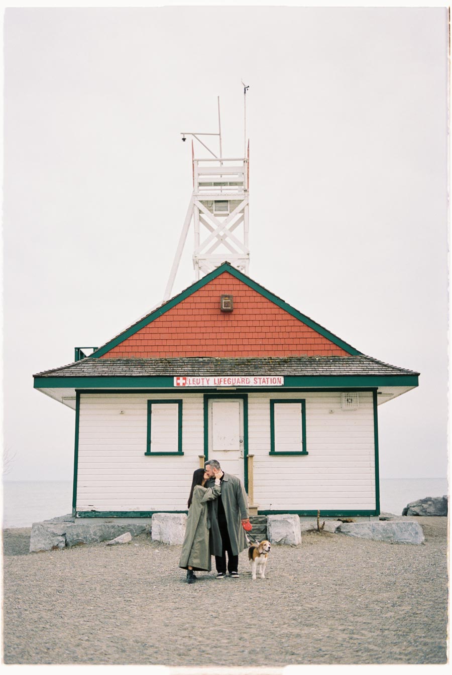 A couple shares a kiss in front of the historic Leuty Lifeguard Station, accompanied by their dog on a cloudy day.