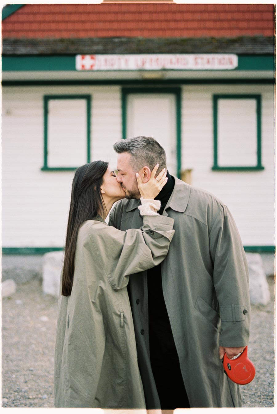 A couple shares a kiss in front of a historic train station, both dressed in long coats, with the man holding a red leash.