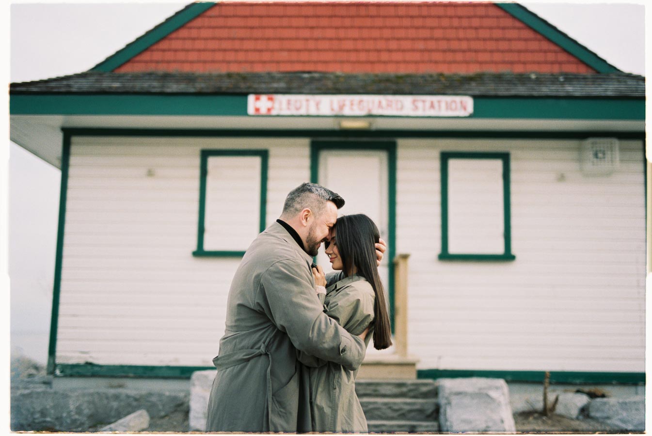 A couple shares a tender moment in front of the Leuty Lifeguard Station, both wearing matching trench coats.