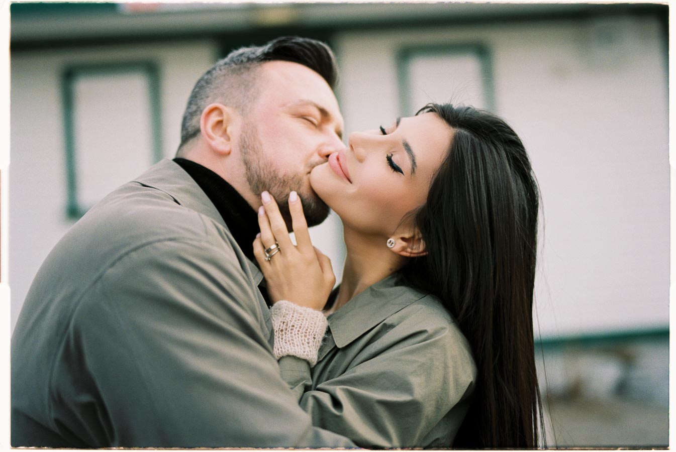 A couple shares a tender moment, embracing and kissing while dressed in matching coats.