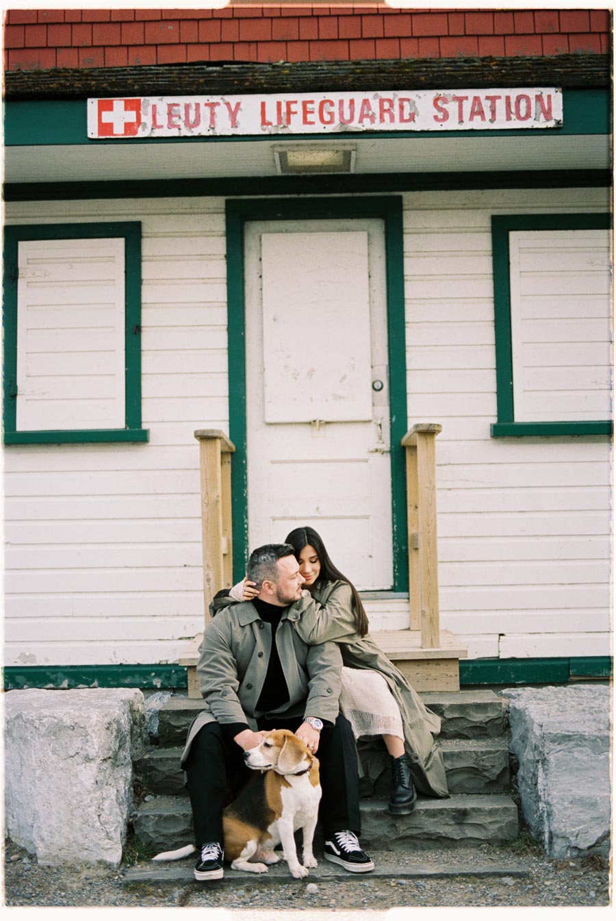 A couple sits affectionately on the steps of the Leuty Lifeguard Station with their dog, enjoying a peaceful moment together.