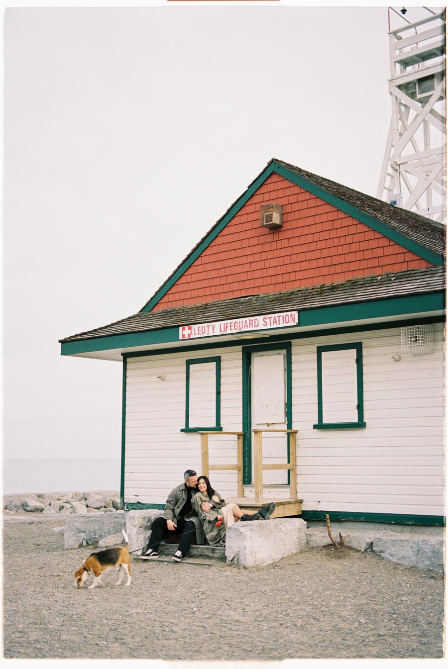 A couple enjoys a moment together on the steps of the Leuty Lifeguard Station, with their dog exploring nearby.