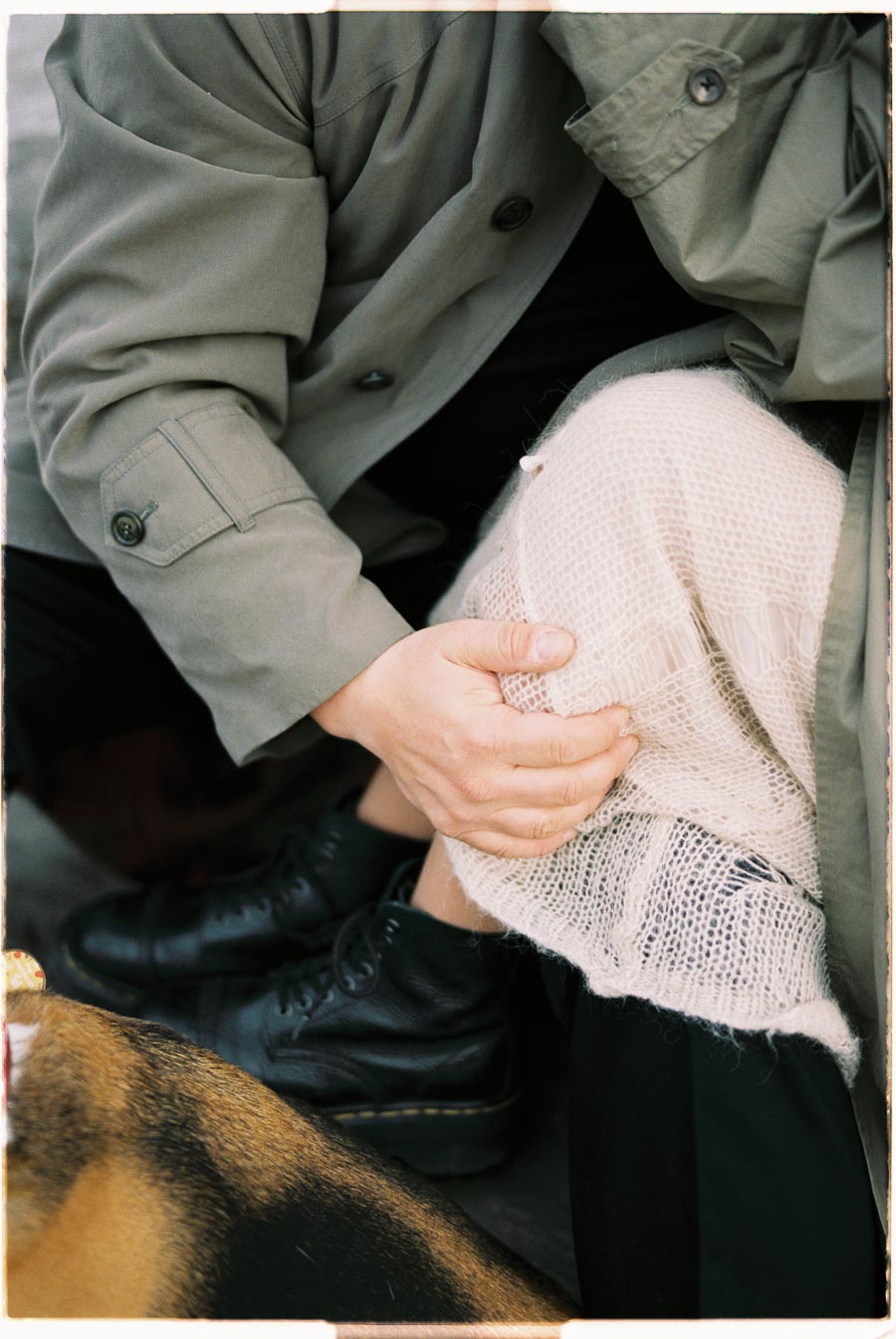 A close-up shot captures a person in a green coat and black boots, gently resting their hand on a knee covered with a white knitted fabric.
