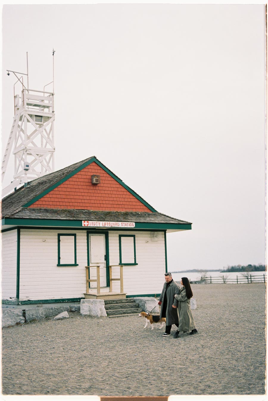 A couple walks their dog near the historic Leuty Lifeguard Station on a sandy beach, under an overcast sky.